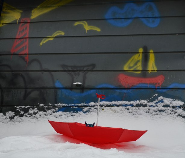 Edgar is in an upside down red umbrella in the snow, in front of a painting on a garage door.  The painting is of a red sailboat sailing beside a whale that is diving under the surface of blue water