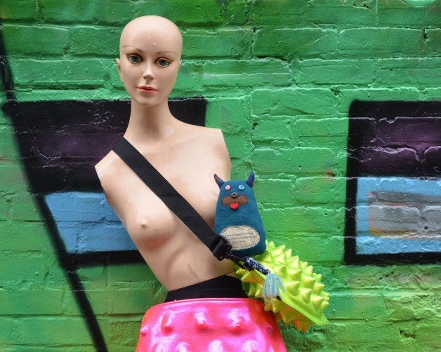 Edgar is with a mannequin that he met in an alley.  She is leaning against a green wall.  She has no hair, and no arms.  She is topless but is wearing a pink skirt and is carry a lime green shoulder bag.  Edgar is sitting on top of the green bag. 
