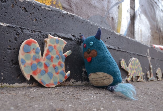 Edgar is standing in an alley.  He is talking to a red and blue spotted cut out in the shape of a squirrel that is about the same size that he is.  A turtle is against the wall behind him and there are a couple of animal shapes in a line behind the turtle. 