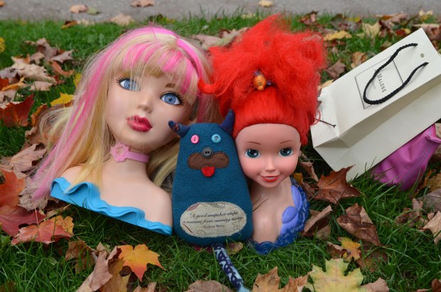 Edgar is sitting between two heads.  One is make-up barbie and the other is Ariel, the little mermaid.  Both of them are bigger than Edgar.  They are on the ground (grass) beside a sidewalk 