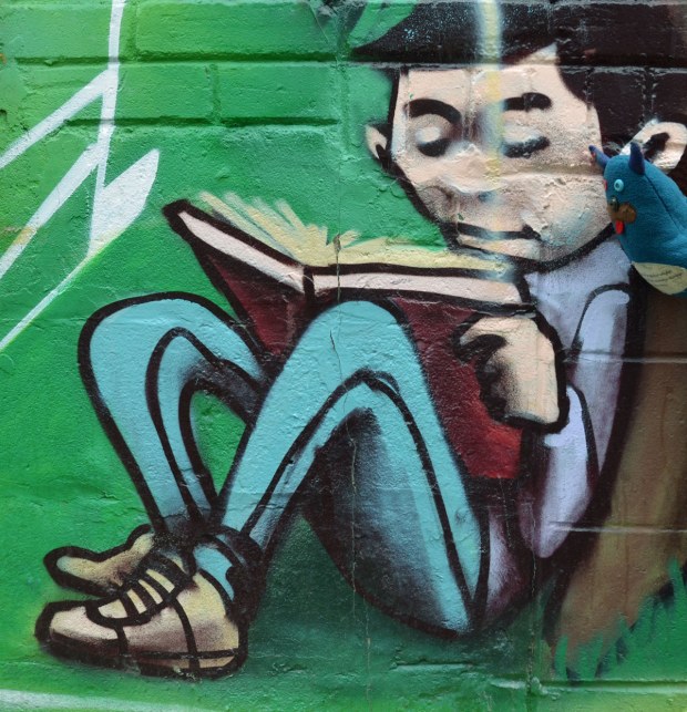 Edgar is reading over the shoulder of a boy, except the boy is actually a painting on a wall and part of a mural.  Green background.  The boy is sitting under a tree with a large book on his knee