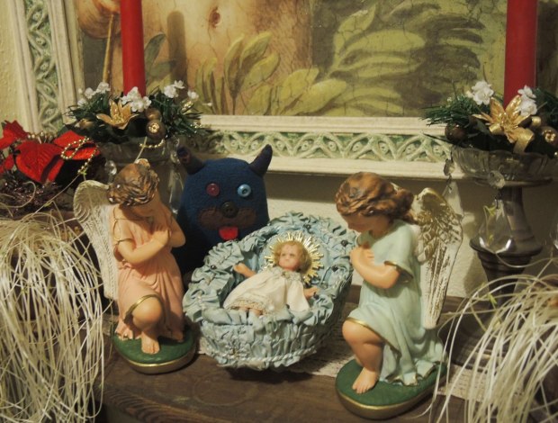 Edgar is standing beside a little ceramic nativity scene.  Baby Jesus is in the manger and two angels are on either side of him.   There are red candles decorated with fake pine and poinsettia