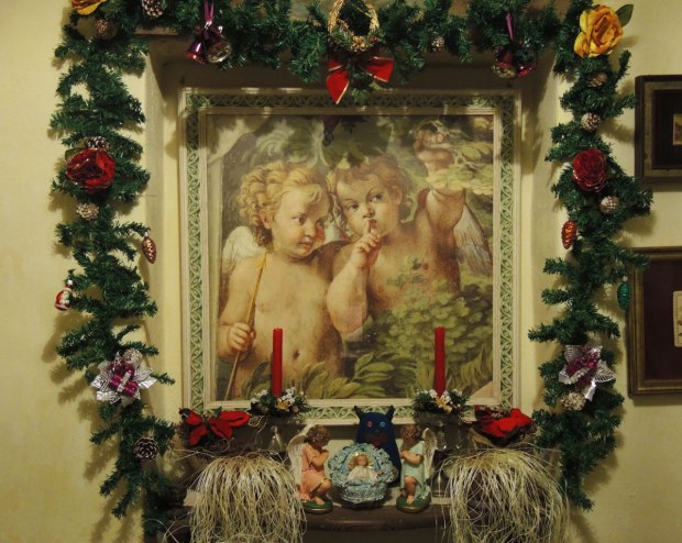 Edgar is standing beside a little ceramic nativity scene.  Baby Jesus is in the manger and two angels are on either side of him.   There are red candles decorated with fake pine and poinsettia.  Above them is a large picture of two little angels in a garden. 