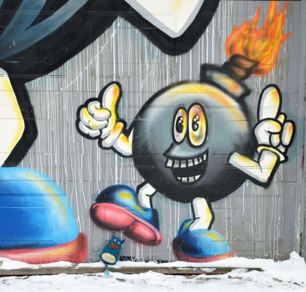 Graffiti painting of a bomb character - a laughing face on the round black bomb, with fire coming out the top, two big white hands and two legs with big blue shoes.  One of the legs is raised and Edgar is sitting under that shoe.  It looks like Edgar is about to get stepped on. 