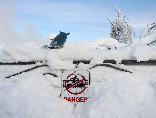 Edgar is standing on top of a snow and ice covered wrought iron fence at Niagara Falls.  There is so much snow you can hardly see the fence.  Some snow has been cleared away to reveal a danger sign that has a picture on it meaning don't climb on or over the fence
