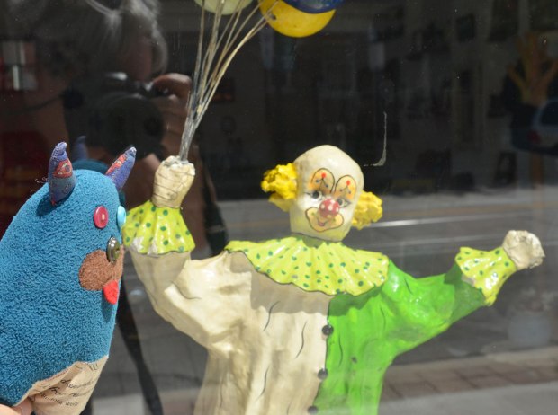 Edgar is looking into a store window.  A figurine of a clown holding a bunch of balloons is looking back at Edgar. 