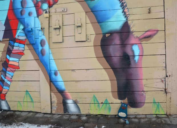 Edgar the little blue monster is sitting on the ground by a mural of a multicoloured horse. It looks like he's right beside the horse's mouth. The horse is standing on four legs but has it's head bent down towards the ground. 