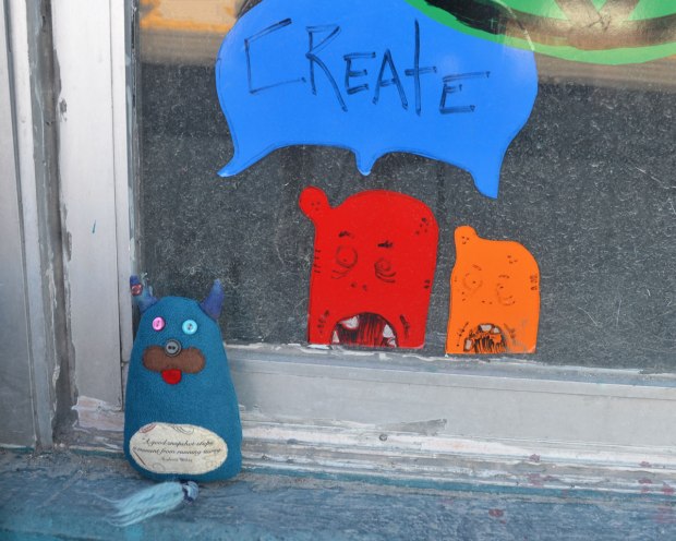 Edgar the little blue monster is sitting by a door with a little red monster and a little orange monster. The word create is in a blue word bubble above their heads so that it looks like they are saying the word together. 