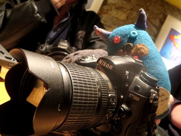 Edgar the little blue monster is checking out the scene as he stands behind a Nikon D7000 DSLR camera. 