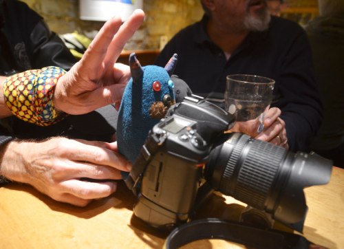 Edgar, the little blue monster, is standing behind a Nikon D7000 DSLR camera on a table. A couple of men are also at the table, one is holding a beer glass beside the camera, one is helping Edgar stand upright and the other is making a peace sign (rabbit ears!) over Edgar's head as the picture is being taken. 