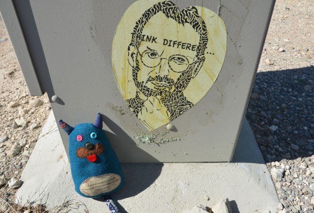 Edgar is sitting beside a grey metal box on a sidewalk. On the box is a wheatpaste picture of a man with beard and glasses with the words think different written on it. 