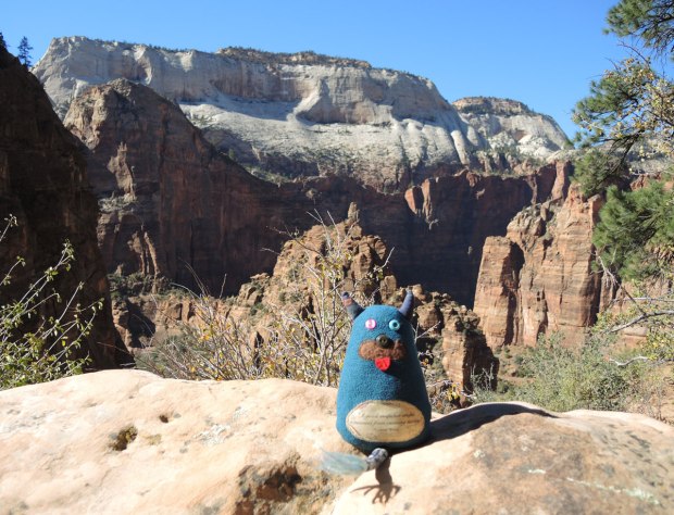 Edgar the little blue monster is sitting on a rock in Zion National Park in Utah. Behind him are more mountains and rocks. 