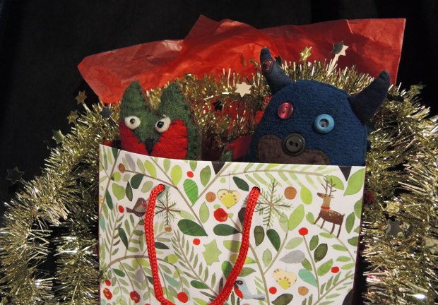 Edgar the little blue monster and his friend Flora are all wrapped up in red tissue paper and a Christmas gift bag and decorated with a shiny gold garland. 