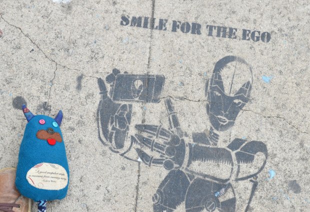 Edgar is lying on the sidewalk beside a stencil of a robot holding a smartphone and taking a selfie. The words with the stencil are smile for the ego 