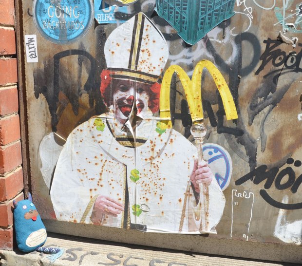 graffiti paste ups on a door in an alley, the pope with the face of Ronald McDonald, plus the yellow M for McDonalds on top of the pope's staff. Edgar is sitting in front it, looking at the pope