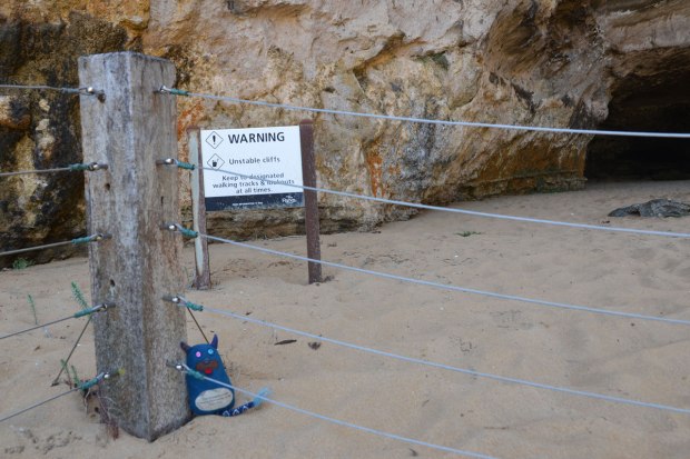Edgar is sitting on the sand, beside a concrete post, and just behind a wire fence. Behind him is a sign that says warning, unstable cliffs, and behind the sign are the cliffs. 