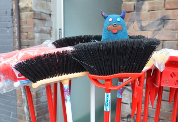 brooms for sale outside a store.  They are brush end up.  Edgar is sitting on one of them. 