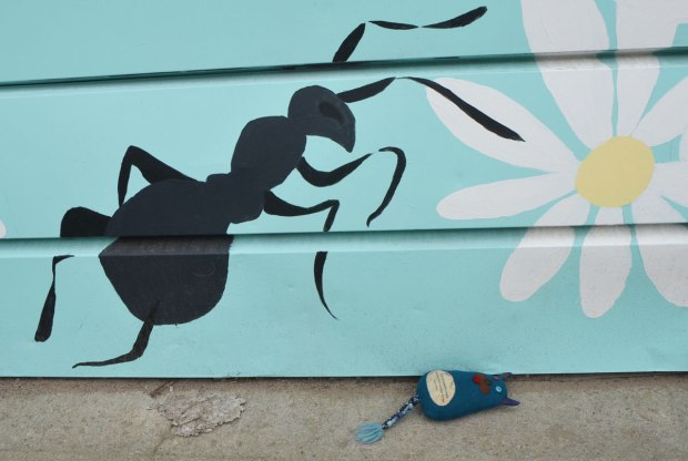 Edgar lies on the pavement beside the bottom of a garage door on which a large black ant has been painted. It looks like the ant is reaching for Edgar