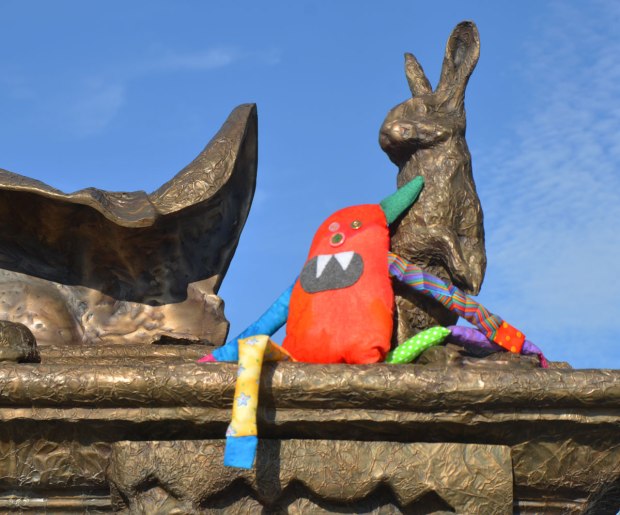 Bo, the rainbow coloured stuffed monster is sitting on a statue beside a small bronze rabbit. 