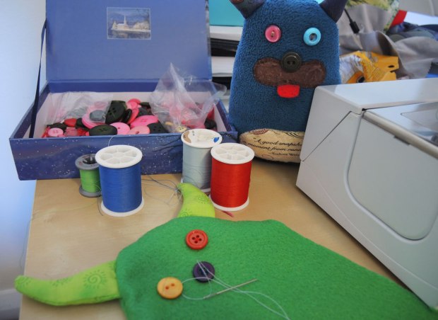 Edgar is standing by a sewing machine, a box of buttons and 4 spools of thread, green, dark blue, light blue and red thread. In the foreground is the beginnings of a new green monster. One red eye has already been sewn on. A yellow eye and a blue nose are in place but haven't been sewn on yet. 