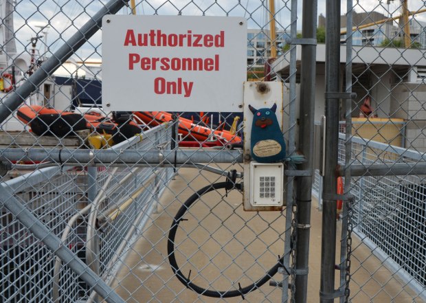 Edgar the little blue monster is standing on top of a keypad that unlocks a chainlink gate. A large sign that says "authorized personnel only" is beside his head. Boats and docks are behind the fence and gate. 