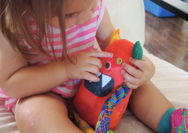 Bo, the rainbow coloured stuffed monster is sitting in a little girl's lap, she is poking at his button eyes 