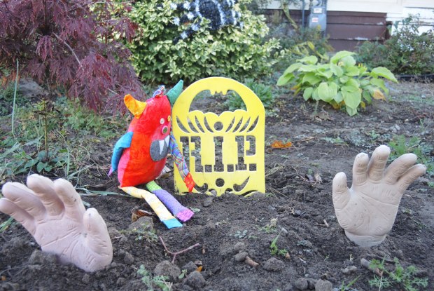 bo, the stuffed monster in many colours, is sitting on the ground beside a yellow RIP tombstone. In front of him two fake hands are coming out of the ground. 
