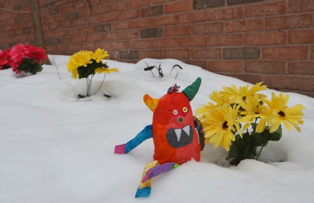 bo the stuffed rainbow coloured monster is sitting in the snow beside a bunch of bright yellow plastic flowers. Other bunches of cloers in reds and purples are also in the photo 