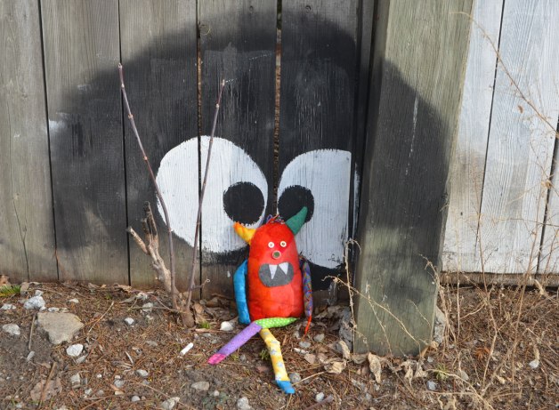 bo, the little rainbow monster is sitting in the dirt on the ground, sitting against a wood fence, a large black circle with two big eyes has been painted on the fence and Bo is sitting between the eyes